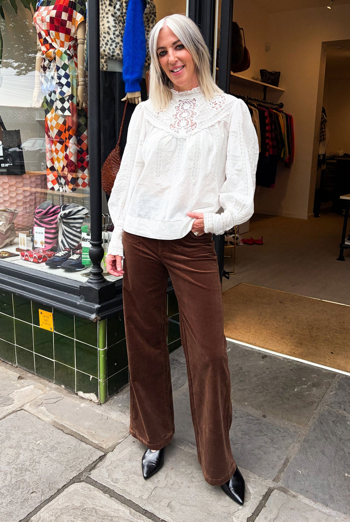 Paige wide leg corduroy trousers in brown, with a white lace-yoke blouse and pointed black boots outside the Kiti shop