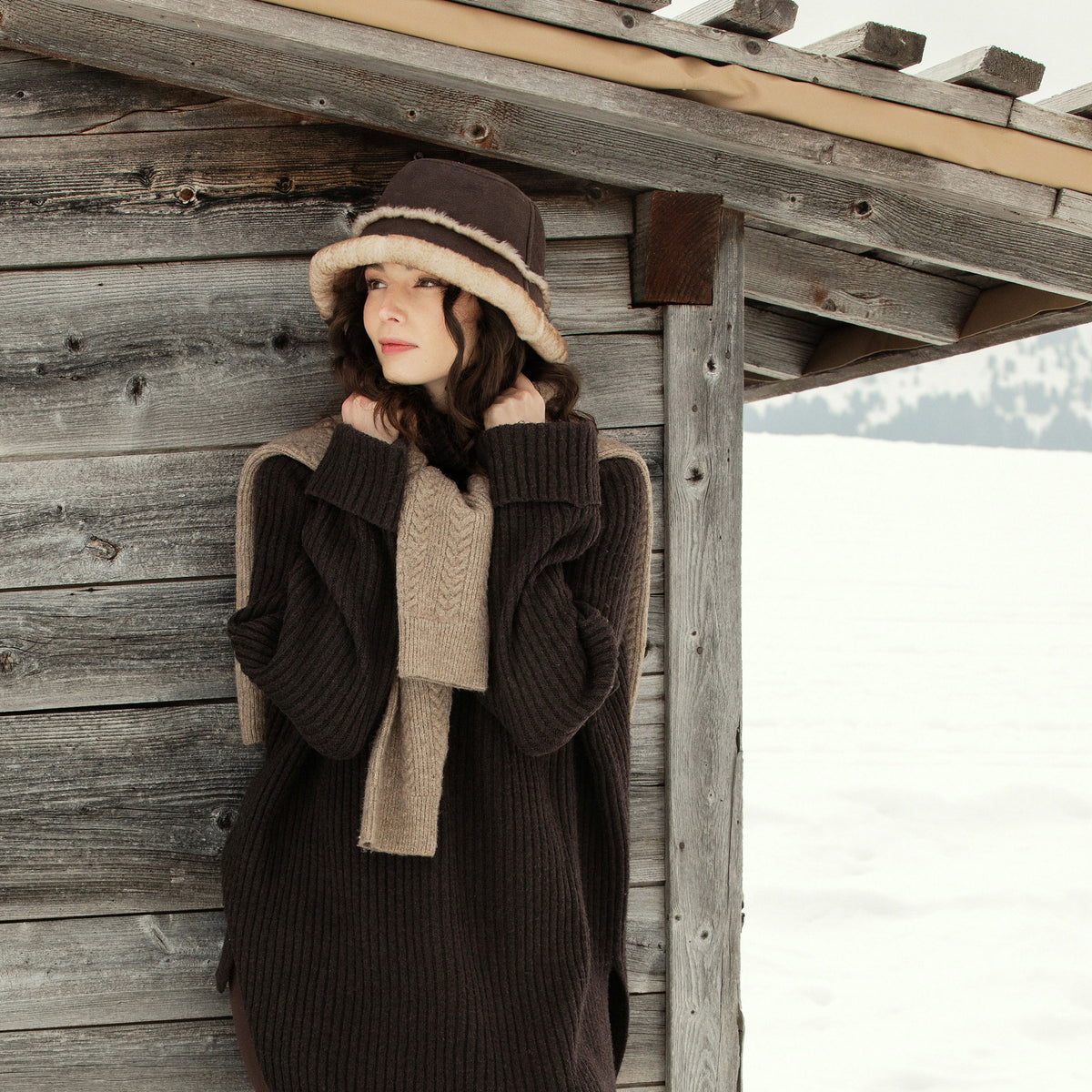 Emu Full-length styling with a chunky ribbed jumper and cable scarf, beside a timber hut in the snow