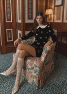 Seated in a floral armchair, long sleeves with frilled cuffs and cream lace knee socks
