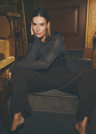Seated in a theatre setting with wide leg dark trousers, sheer sleeves catching the light